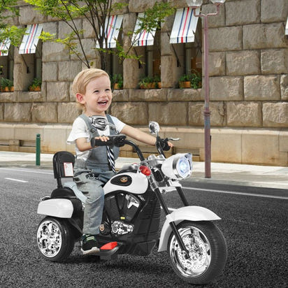 Child riding a toy motorcycle on a city street with stone buildings and greenery in the background.